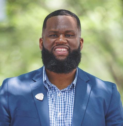 A man in a blue blazer and dress shirt stands against a backdrop of trees.